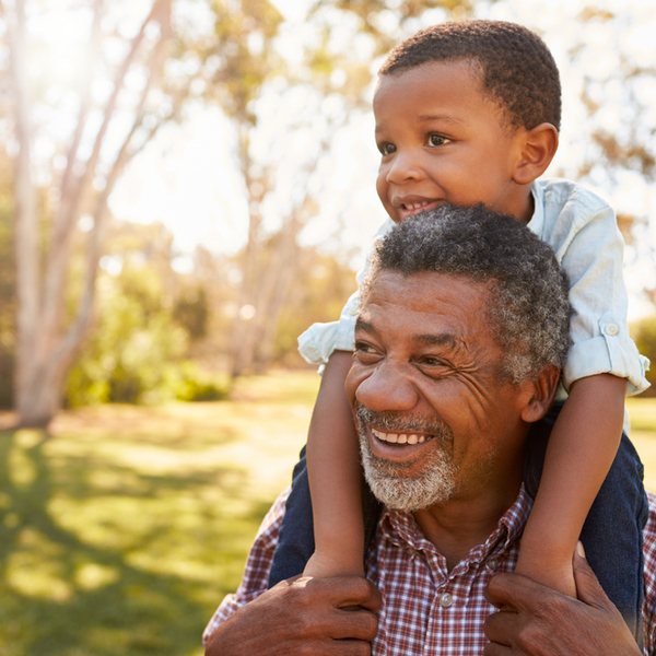 A grandfather carrying his grandson on his shoulders through a park.