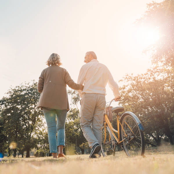 An older couple walking through a park as one pushes a bike.