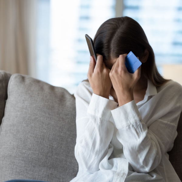 A woman holds her head with a phone in one hand and a bank card in the other.